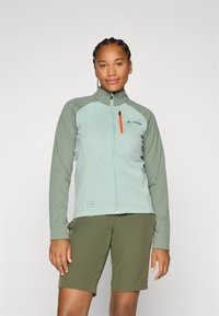 En kvinne med fletter har på seg en lys grønn jakke og mørkegrønne shorts, og smiler selvsikkert mot en enkel grå bakgrunn.