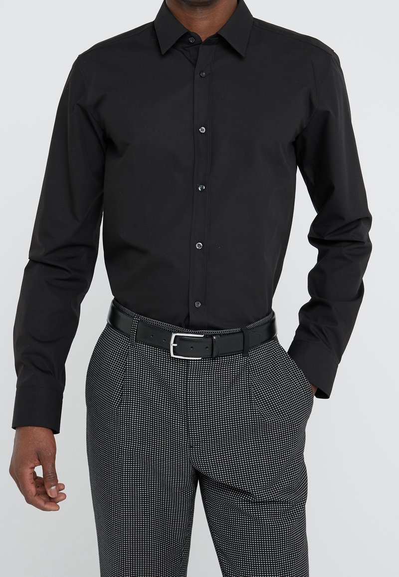 Chemise noire à manches longues avec un col pointu et une ouverture boutonnée sur le devant. Portée avec un pantalon gris à motif et une ceinture noire avec une boucle argentée.