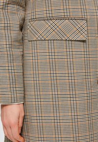 Nærbillede af en beige- og brunternet jakke lomme med låg, som viser en persons venstre hånd, der hviler ved siden af jakken.