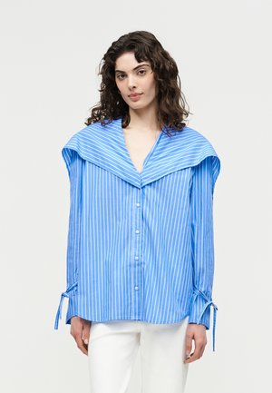 Žena s kučeravými vlasmi má na sebe modro-bielu vertikálne prúžkovanú oversized košeľu so širokým golierom a šnúrkami na rukávoch, v kombinácii s bielymi nohavicami.