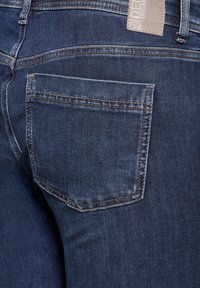 Jean en denim bleu foncé avec une poche arrière. Le tissu a une texture lisse avec des coutures visibles et une étiquette à la taille.