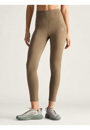 Woman wearing high-waisted brown leggings and beige-gray sneakers, standing on a plain light background.