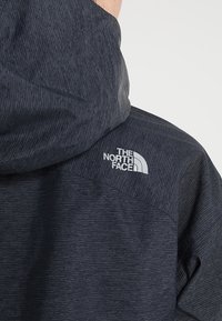 Personne portant une veste d'extérieur texturée gris foncé avec une capuche et le logo "The North Face" sur le haut du dos.