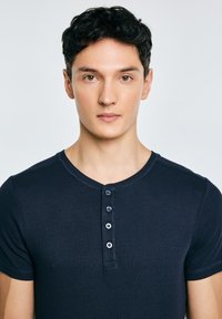 Młody mężczyzna z krótkimi kręconymi ciemnymi włosami, ubrany w ciemnoniebieską koszulkę henley z krótkim rękawem, zwrócony przodem na jasnym, jednolitym tle.