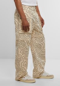 Beige cargo pants with contrasting black wavy line patterns, featuring side pockets and a relaxed fit, paired with white sneakers.