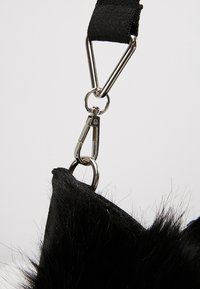 Zwart, harig textiel bevestigd aan een metalen karabijnhaak en driehoekige ring, verbonden met een zwarte riem tegen een lichte achtergrond.