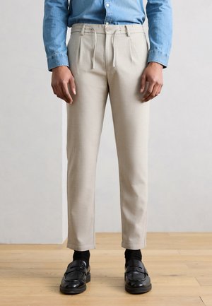 Person wearing beige trousers with front pleats, black socks, black loafers, and a tucked-in blue denim shirt, standing on a wooden floor.