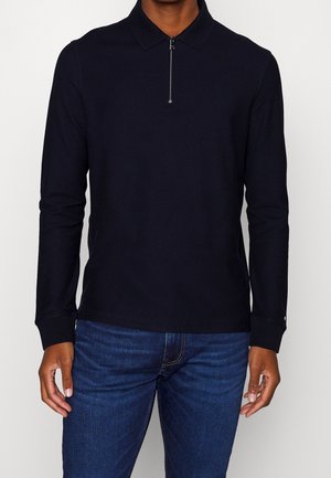 Homme portant un polo noir uni à manches longues avec une fermeture éclair frontale, associé à un jean en denim bleu, debout devant un fond uni.