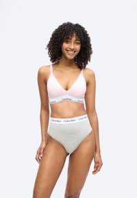 Χαμογελαστή γυναίκα με σγουρά μαλλιά που φοράει ένα ανοιχτό ροζ σουτιέν Calvin Klein και γκρι εσώρουχα Calvin Klein με υψηλή μέση, σε λευκό φόντο.