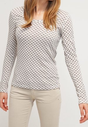 Vrouw draagt een wit shirt met lange mouwen met een klein grijs ruitjespatroon en beige broek tegen een effen achtergrond.