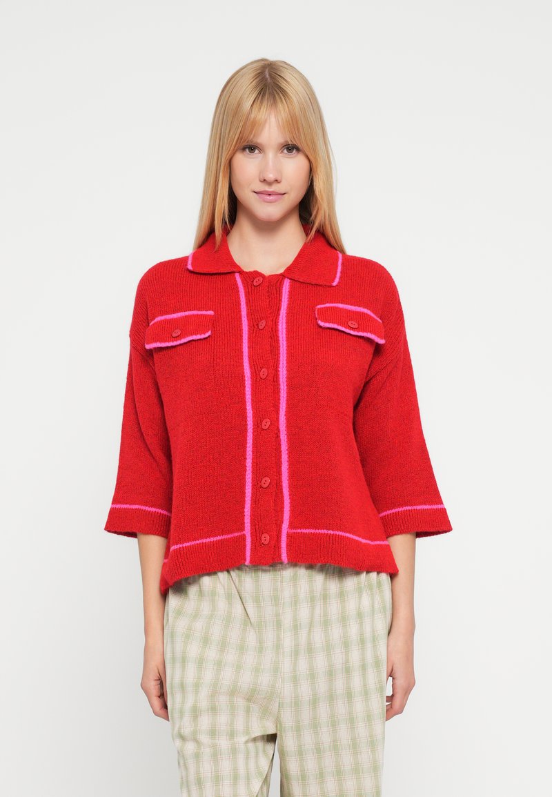 Sarkans adīts džemperis ar rozā akcentiem, ar apkakli, pogām priekšā un divām krūšu kabatām. Platas piedurknes un teksturēts audums. Kombinēts ar rūtainām biksēm.