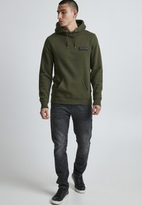 Olive green hoodie made of cotton fabric, featuring a front pocket, drawstring hood, and a small logo patch. Worn with faded black jeans.
