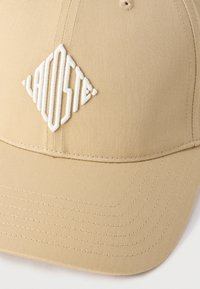 Beežikarva baseballi nokamüts tekstureeritud kangaga, valge tikandiga teemantkujulise logoga ning detailse õmblusega mütsi ääres.