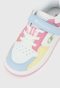 Λευκό αθλητικό παπούτσι με ροζ, μπλε και κίτρινες επιφάνειες, λευκά κορδόνια, μπλε λουράκι με το λογότυπο της Champion και τρύπες στην περιοχή των δαχτύλων.