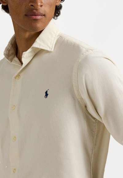 Polo de culoare bej, fabricat dintr-un material texturat, având un guler pliat și un logo brodat albastru pe pieptul stâng. Închidere cu nasturi.