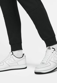 Valkoiset Nike Air Force 1 -tennarit mustilla yksityiskohdilla, joissa on rei'itetty varvas, litteät nauhat ja teksturoitu kumipohja. Mustat verkkarit näkyvissä.