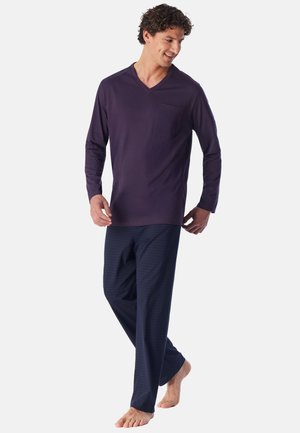 Mann trägt ein dunkelviolettes langärmeliges V-Ausschnitt-Shirt mit Tasche und lockere marineblaue Hose, geht barfuß vor einem schlichten hellen Hintergrund.