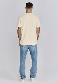 Camiseta de algodón crema con mangas cortas y corte recto, combinada con jeans de denim de corte relajado en azul claro, llevada con zapatillas blancas.