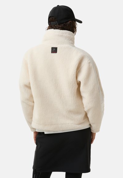 Fløjljakke i cremefarve med høj krave, tekstureret overflade og et firkantet mærke på ryggen. Kombineret med et sort nederdel.