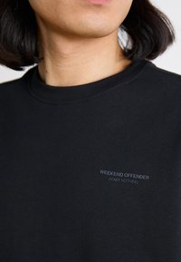 T-shirt in cotone nero con colletto rotondo, maniche corte e un piccolo logo ricamato bianco sul lato sinistro del petto con la scritta "WEEKEND OFFENDER ADMIT NOTHING."