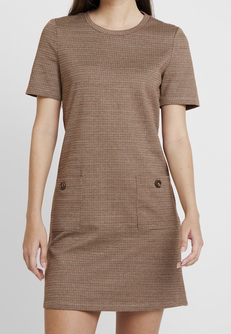 Femme portant une mini-robe à manches courtes, à carreaux marron et beige, avec un col rond et deux poches plaquées avant avec boutons.