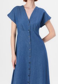 Sininen farkkutunika, jossa on lyhyet hihattomat hihat, V-pääntie, etupainikkeet ja kerääntynyt vyötärö, jossa on virtaava A-linjainen siluetti.