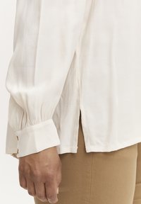 Blusa beige a maniche lunghe con vestibilità rilassata, caratterizzata da un polsino maniche plissettato e spacchi laterali all'orlo, realizzata in un tessuto leggero e testurizzato.