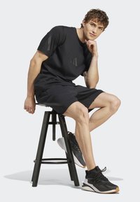 Melnā sporta kreklā ar smalku tonālo akcentu, apvienots ar melnām šortiem un melniem sporta apaviem. Modelis pozē uz melnas taburetes.