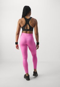 Svart sportbh med racerback-design, tillsammans med figurnära rosa leggings. Svarta träningsskor och handledsband synliga.