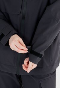 Svart vattentät jacka med dragkedja fram och justerbara kardborreband runt ärmarna. Har en jämn yta och en minimalistisk design.