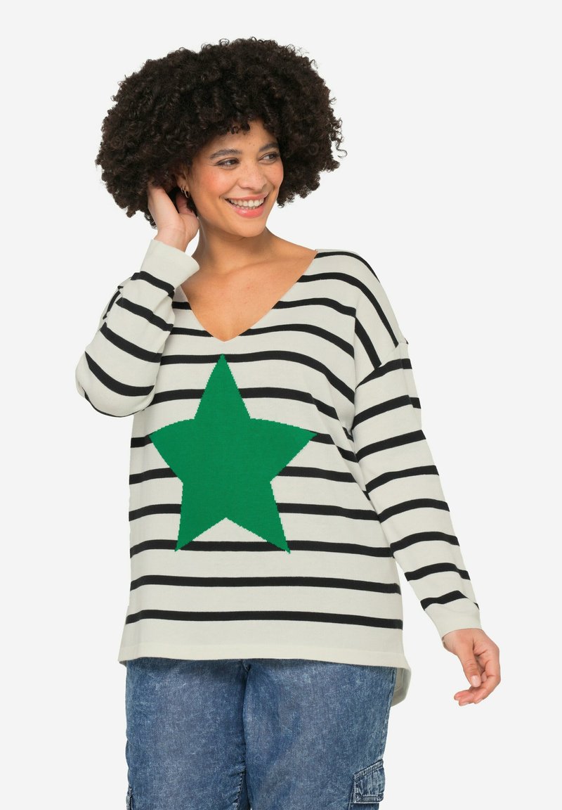 Sweter w paski w kolorze off-white, z dużą zieloną gwiazdą na środku. Głęboki dekolt w serek i luźny krój z długimi rękawami. Widoczne dżinsy.
