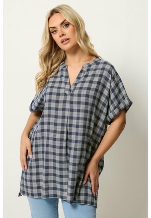Blonde vrouw die een losse marineblauwe en witte geruite tuniek met korte mouwen en lichtblauwe spijkerbroek draagt, poserend met handen in de zij.