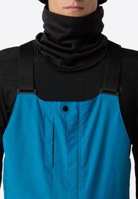 Man in felblauwe tuinbroek met zwarte bandjes en een zwarte nekwarmer over een zwart shirt met lange mouwen.