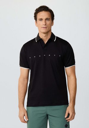 Mann trägt schwarzes Poloshirt mit weißem Rand und "Hackett"-Schriftzug, steht vor einem schlichten hellen Hintergrund.