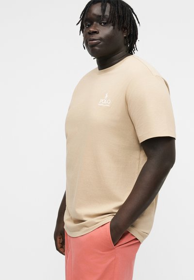 Ung mann med mørk hud og dreadlocks, iført en beige Polo Ralph Lauren t-skjorte og korallrosa bukser, hendene i lommer, ser fremover.