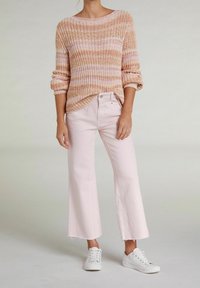 Donna che indossa un maglione a righe rosa e beige, pantaloni a taglio largo di un tenue rosa e sneakers bianche, in piedi su uno sfondo semplice.