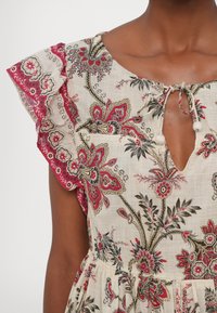 Robe à motif floral avec des couleurs rouge, noir et beige, manches volantes, encolure en trou de serrure et fermeture par lien avec pompons.