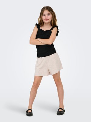 Girl standing with arms crossed wearing a black sleeveless top, beige shorts, and black shoes on a plain light background.
