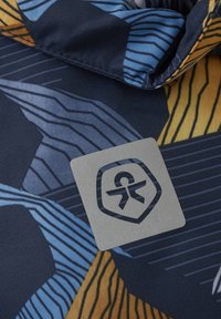 Mönstrad tyg med blå, gul och marinblå nyanser, med en texturerad yta. Inkluderar en fyrkantig, silverfärgad logotagg med ett emblem.