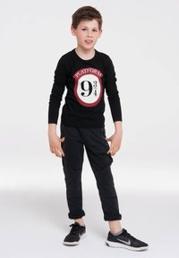 Schwarzes Langarmshirt mit rotem und weißem "Platform 9¾"-Graphic, kombiniert mit schwarzen strukturierten Hosen und schwarzen Sportschuhen.