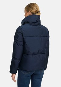 Vrouw in een marineblauwe gewatteerde jas met een hoge kraag, gezien van achteren, gecombineerd met blauwe jeans.