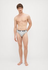 Hallid puuvillased aluspüksid musta Moschino logo vööga, millel on sobituv disain ja sile tekstuur. Mudel seisab paljajalu, käed külgedel.