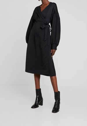 Femme portant une robe portefeuille noire à manches longues bouffantes et des bottines à talons noires, debout devant un fond gris uni.