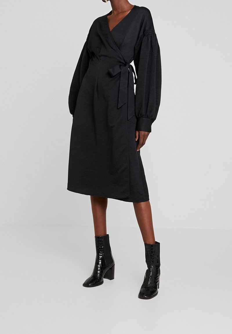 Femme portant une robe portefeuille noire à manches longues bouffantes et des bottines à talons noires, debout devant un fond gris uni.