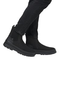 Bottines noires avec une finition lisse, des panneaux latéraux élastiques et une semelle épaisse et texturée. Portées avec un jean sombre.