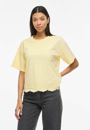 Femme aux longs cheveux noirs portant un haut ample à manches courtes de couleur jaune pâle avec un ourlet festonné et un jean gris foncé, debout avec une main sur la hanche.