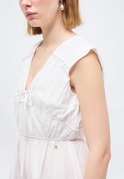 Jeune femme aux cheveux châtain clair portant une robe blanche sans manches avec un nœud à l'avant et une boucle d'oreille argentée en forme de goutte, sur fond uni.