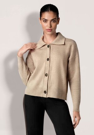Femme en cardigan beige boutonné avec un col large et un pantalon noir, debout devant un fond clair uni.