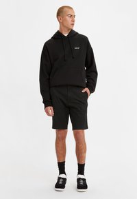 Svart hettegenser med frontlomme og snor, kombinert med svarte shorts. Har en logo på brystet, brukt med svarte joggesko og sokker.