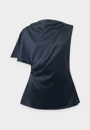 Femme aux longs cheveux foncés portant un haut sans manches drapé bleu marine et un pantalon large beige, debout devant un fond clair uni.